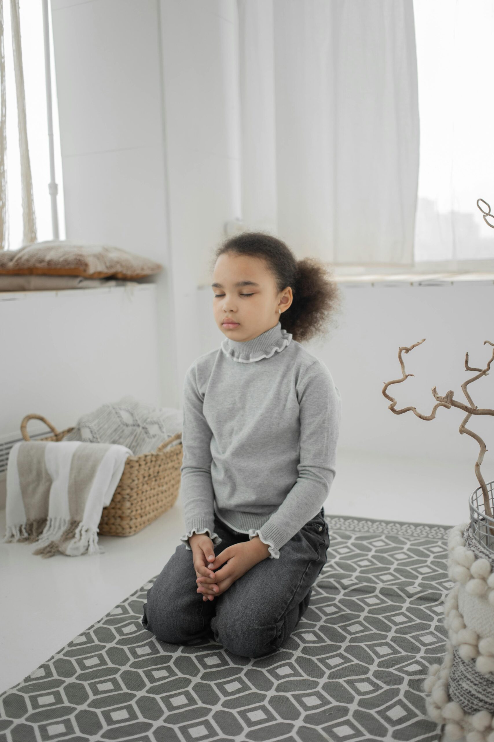 Full body of calm little African American girl with curly hair in casual clothes sitting on floor and meditating with closed eyes at home